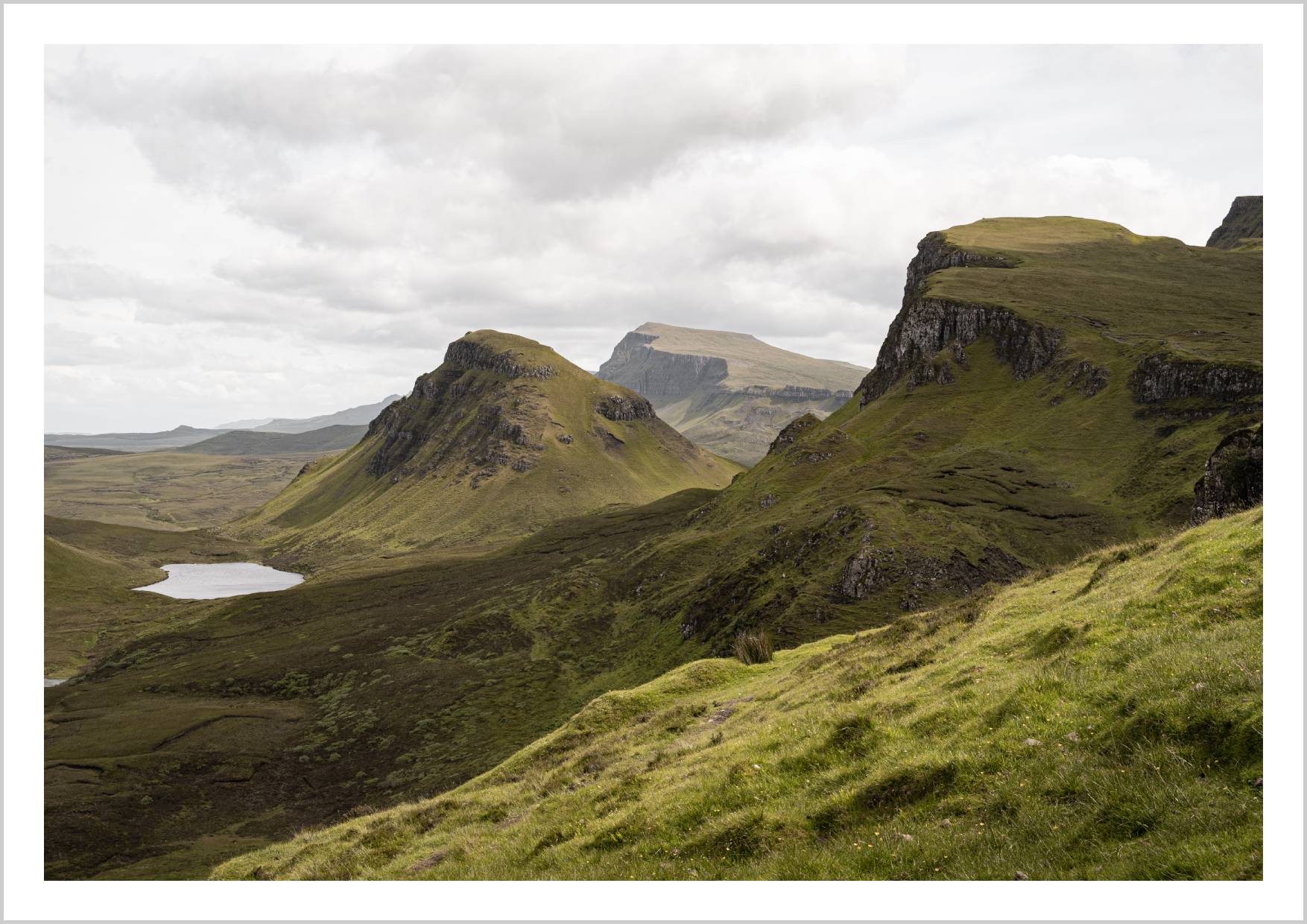 A panoramic view of the Quiraing landscape on the Isle of Skye, featuring green hills and rocky cliffs under a cloudy sky. – Arteve Gallery