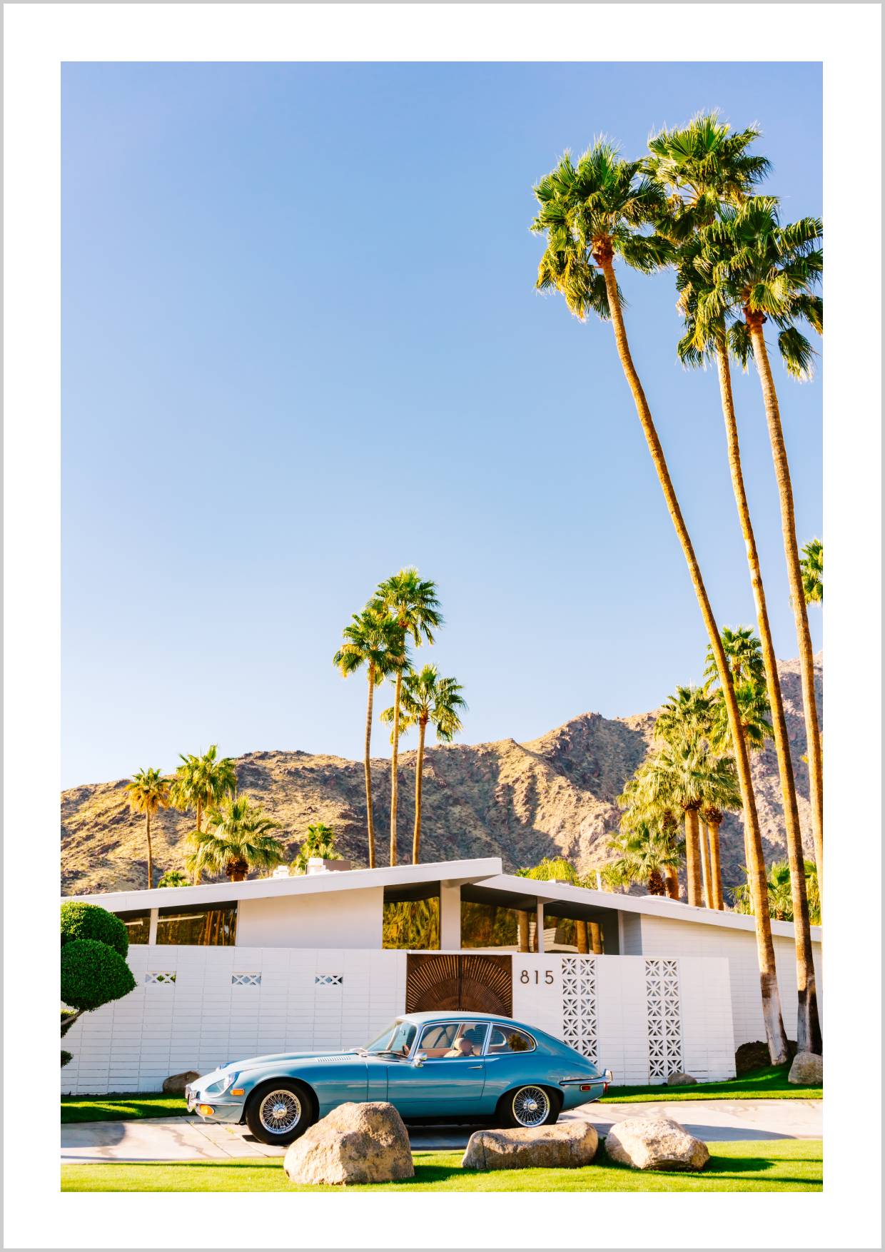 A vintage blue car parked near a modern white house with palm trees and mountains in the background. – Arteve Gallery