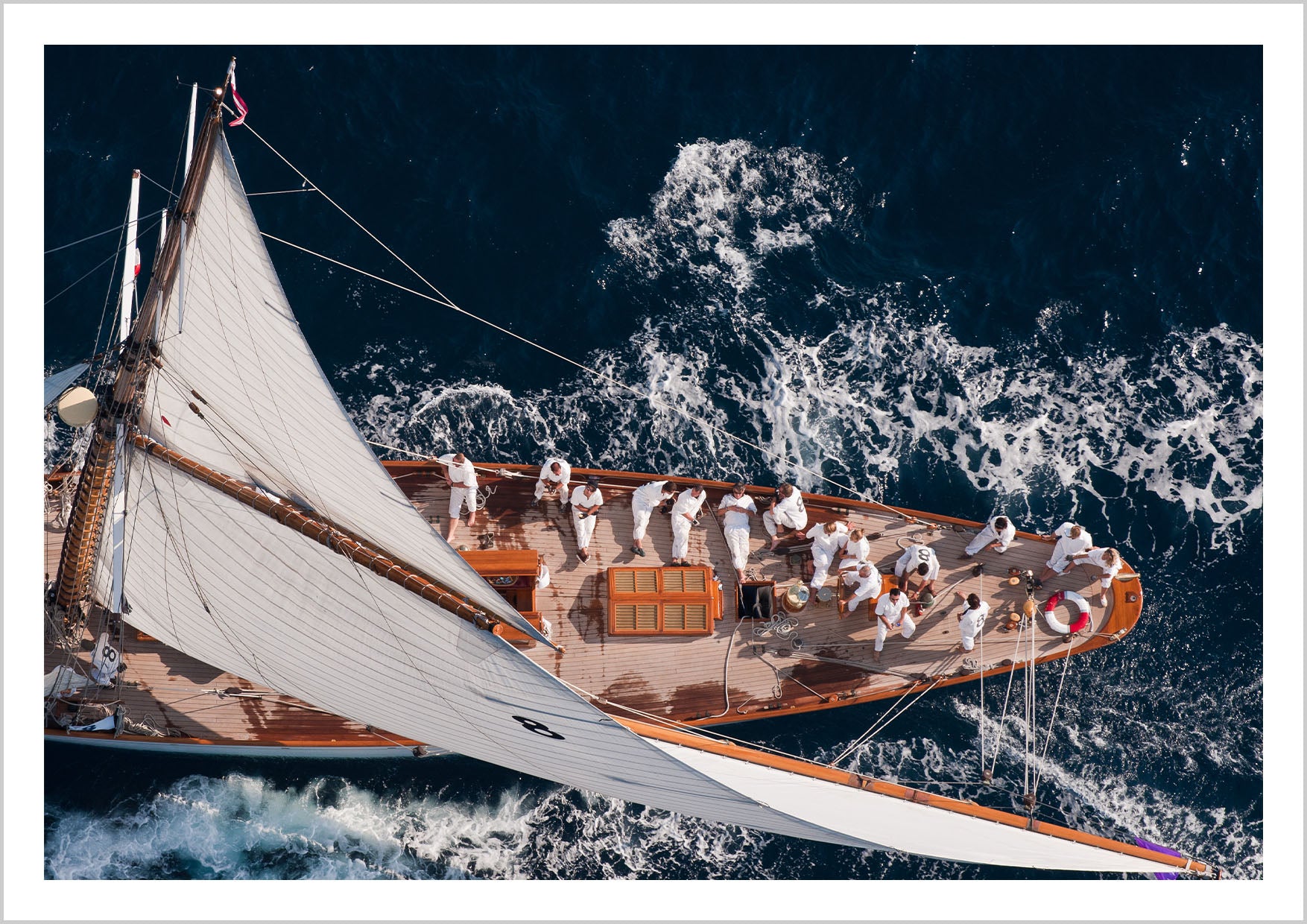 An aerial view of a sailboat with sailors on deck, navigating through deep blue waters. – Arteve Gallery