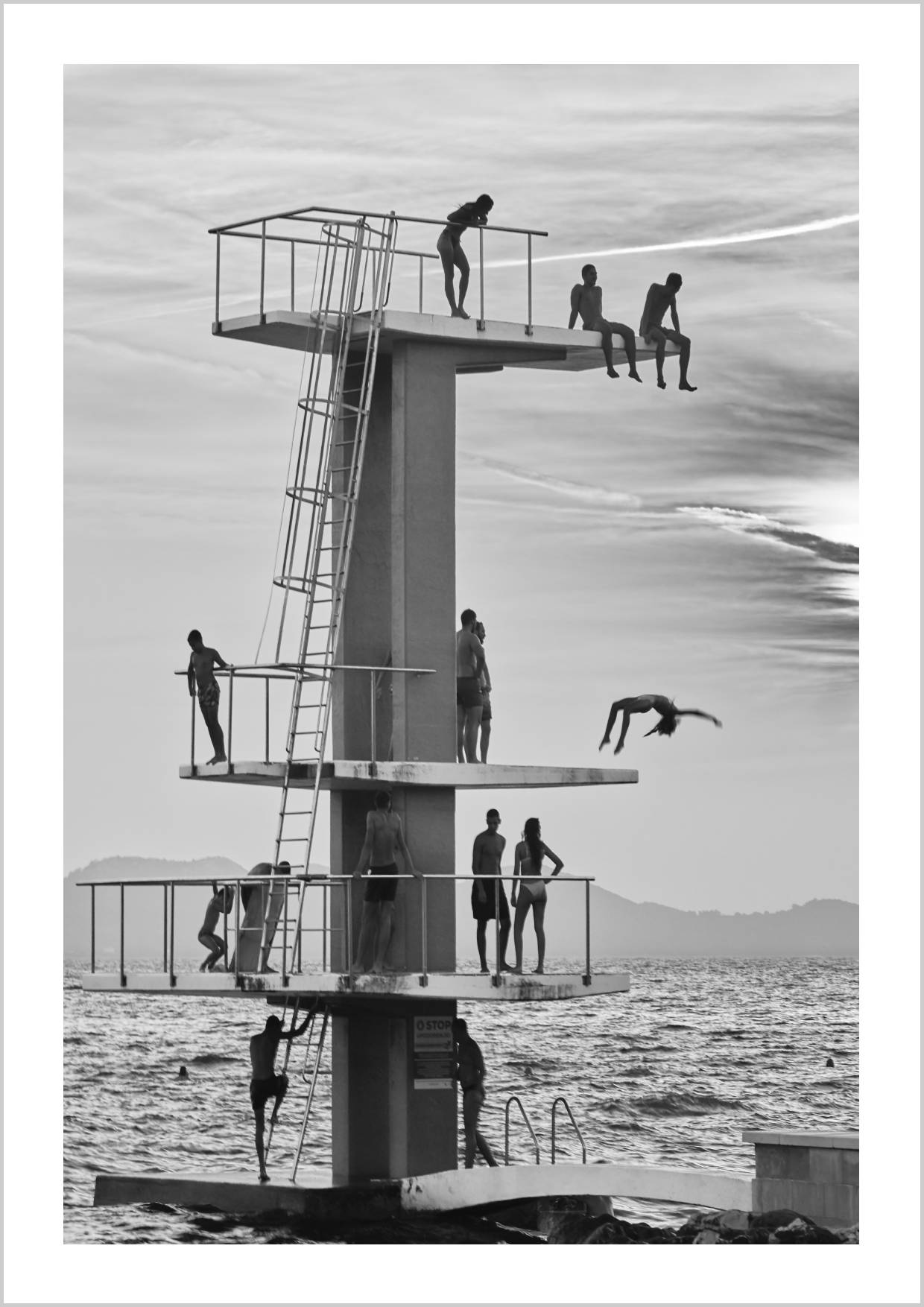 Monochrome image of a diving tower with people diving and sitting on platforms by the ocean. – Arteve Gallery