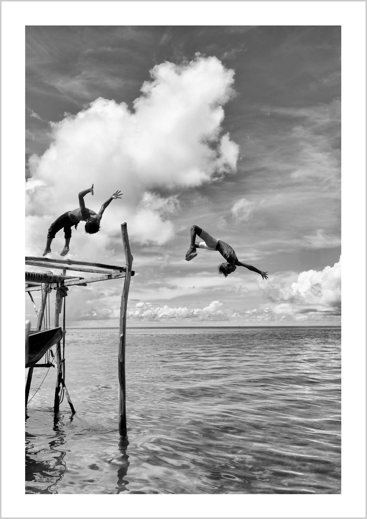Two children leaping off a wooden structure into the ocean, surrounded by clouds. – Arteve Gallery
