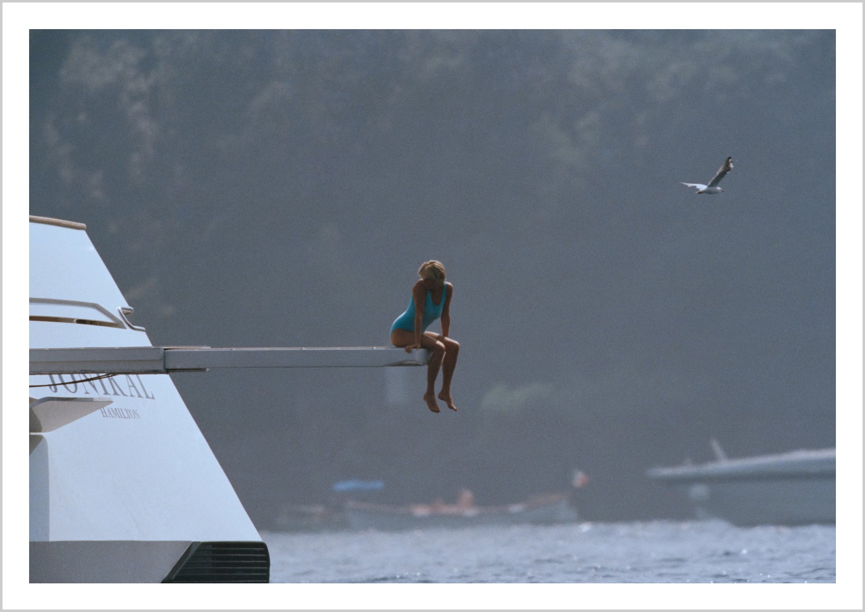 A person in a blue swimsuit sitting on the edge of a boat, gazing into the distance, with soft blue and gray tones in the background. – Arteve Gallery