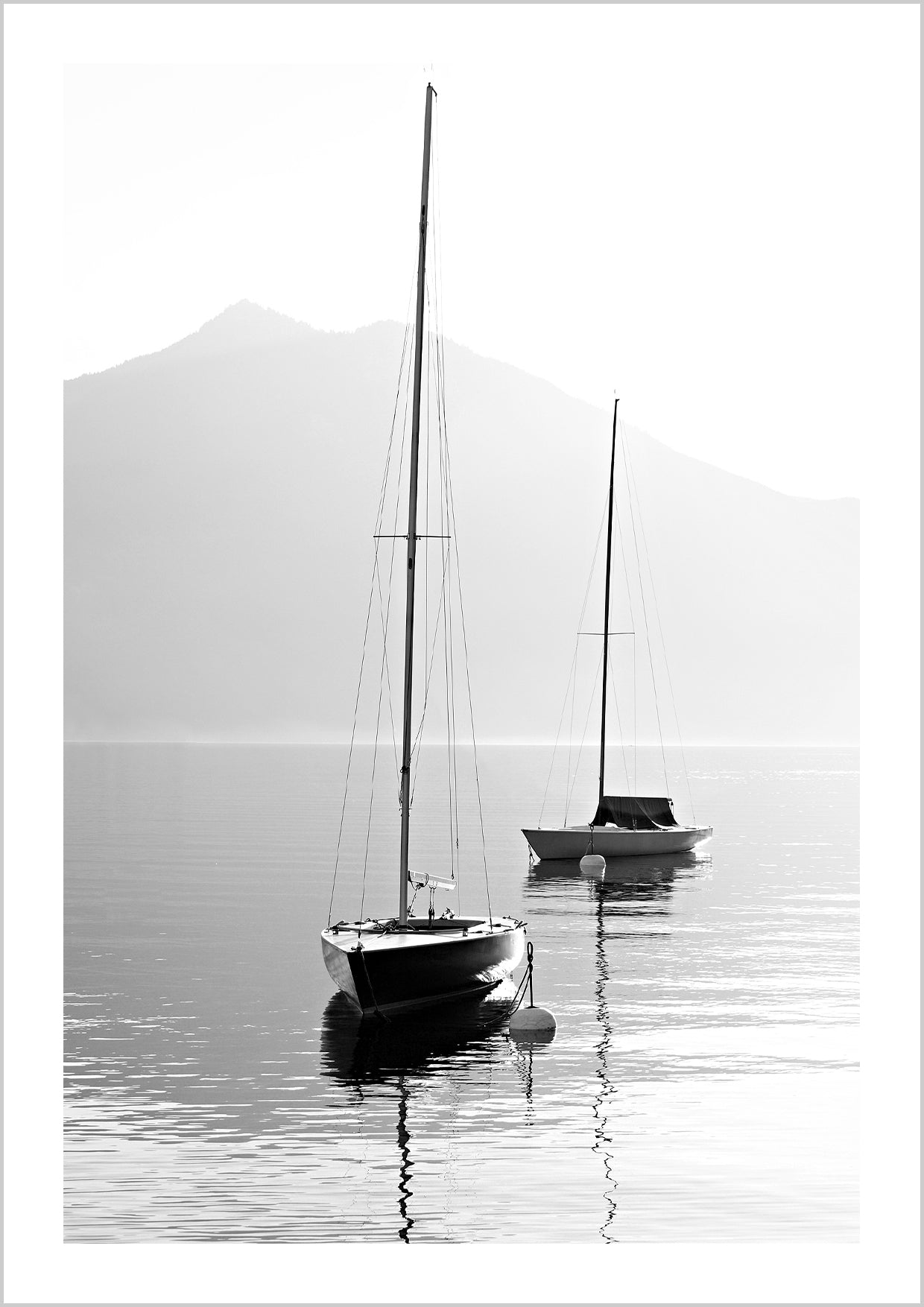 Harmonic photo poster in black and white with motifs of two sailboats floating peacefully on a calm sea. In the background, the silhouette of an angular mountain stretches toward the grey sky. 
