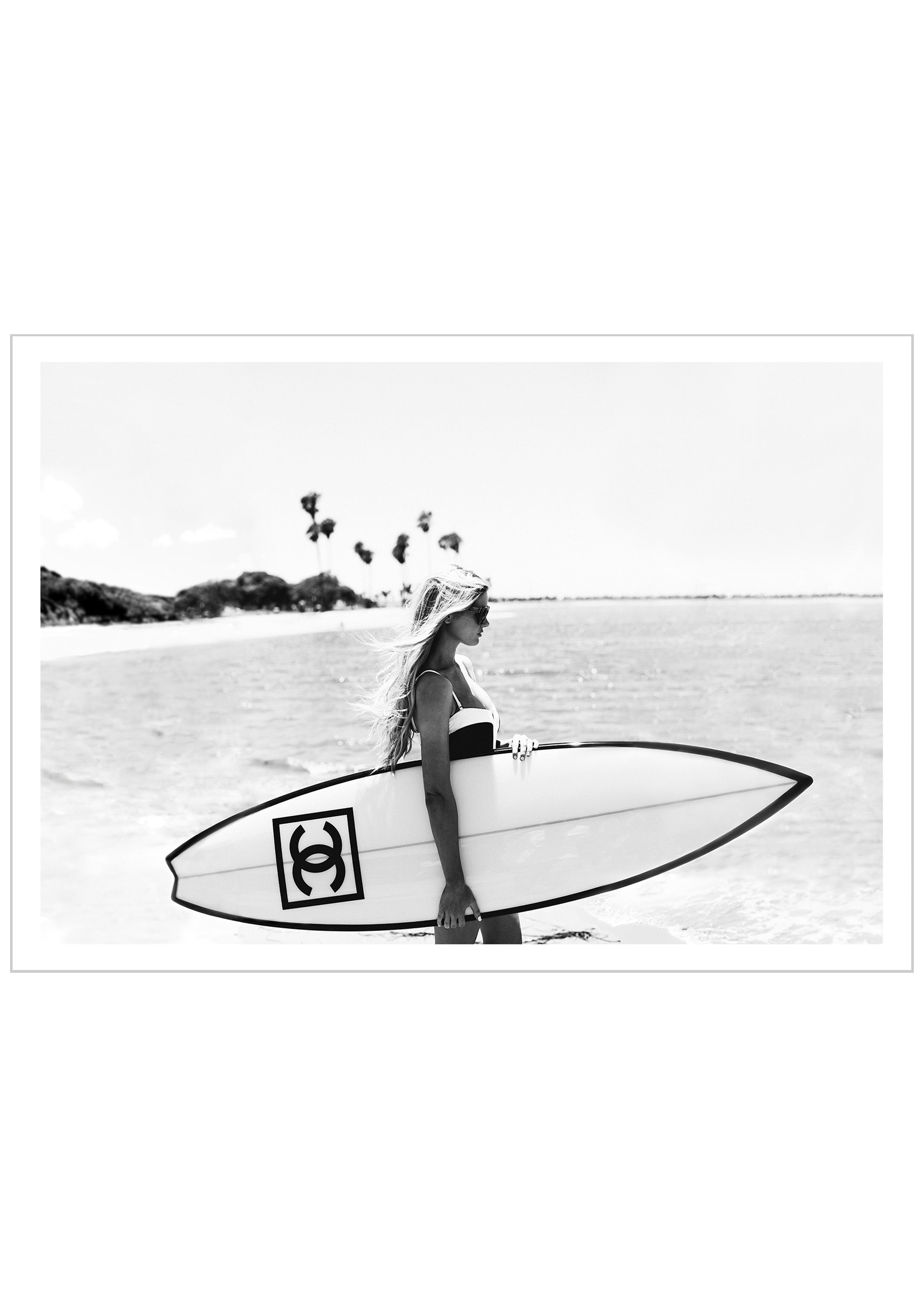 Photograph of a bikini girl holding a Chanel surfboard toward the sea.