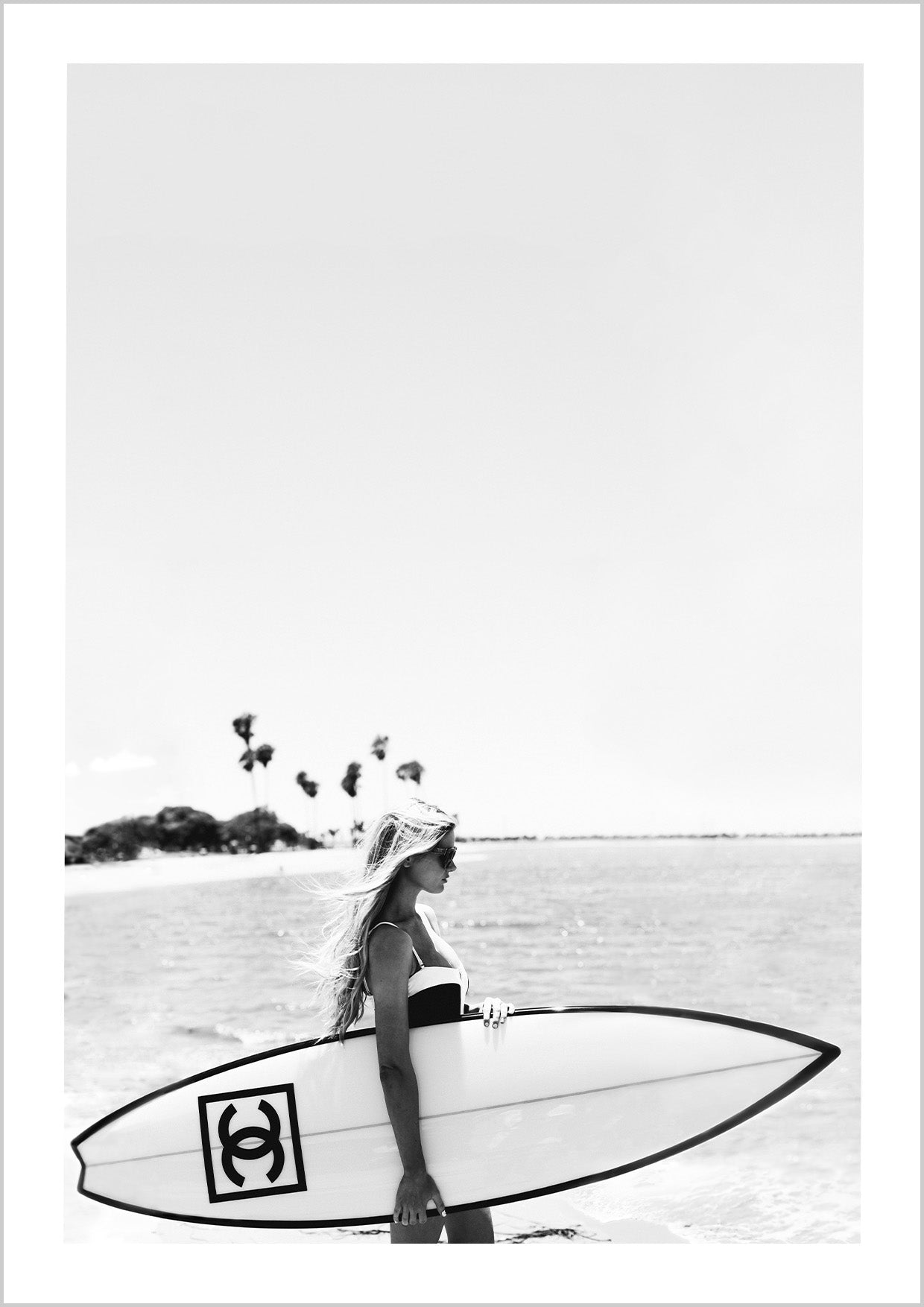 Photograph of a bikini girl holding a Chanel surfboard toward the sea. 