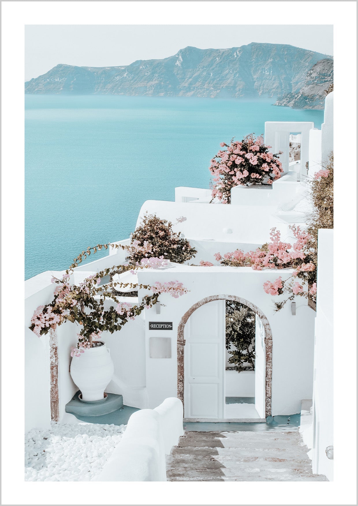 A beautiful landscape poster with a view of the Greek island of Santorini and the crystal clear blue sea. The traditional white house facades are decorated with pink flowers and make the Poster radiate.