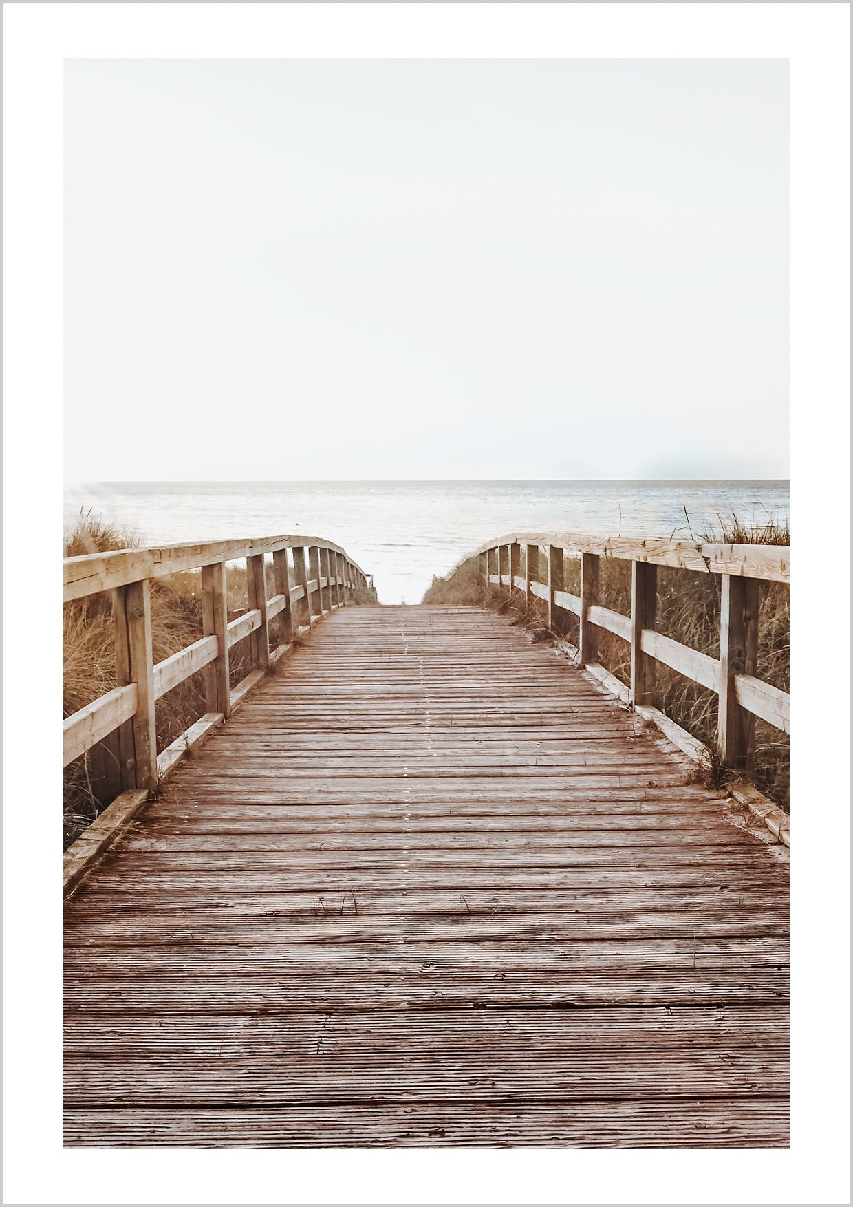 Photograph of a wooden path leads down to a beach and the calm ocean. The gentle sun rays give the photographic Poster a warm atmosphere.