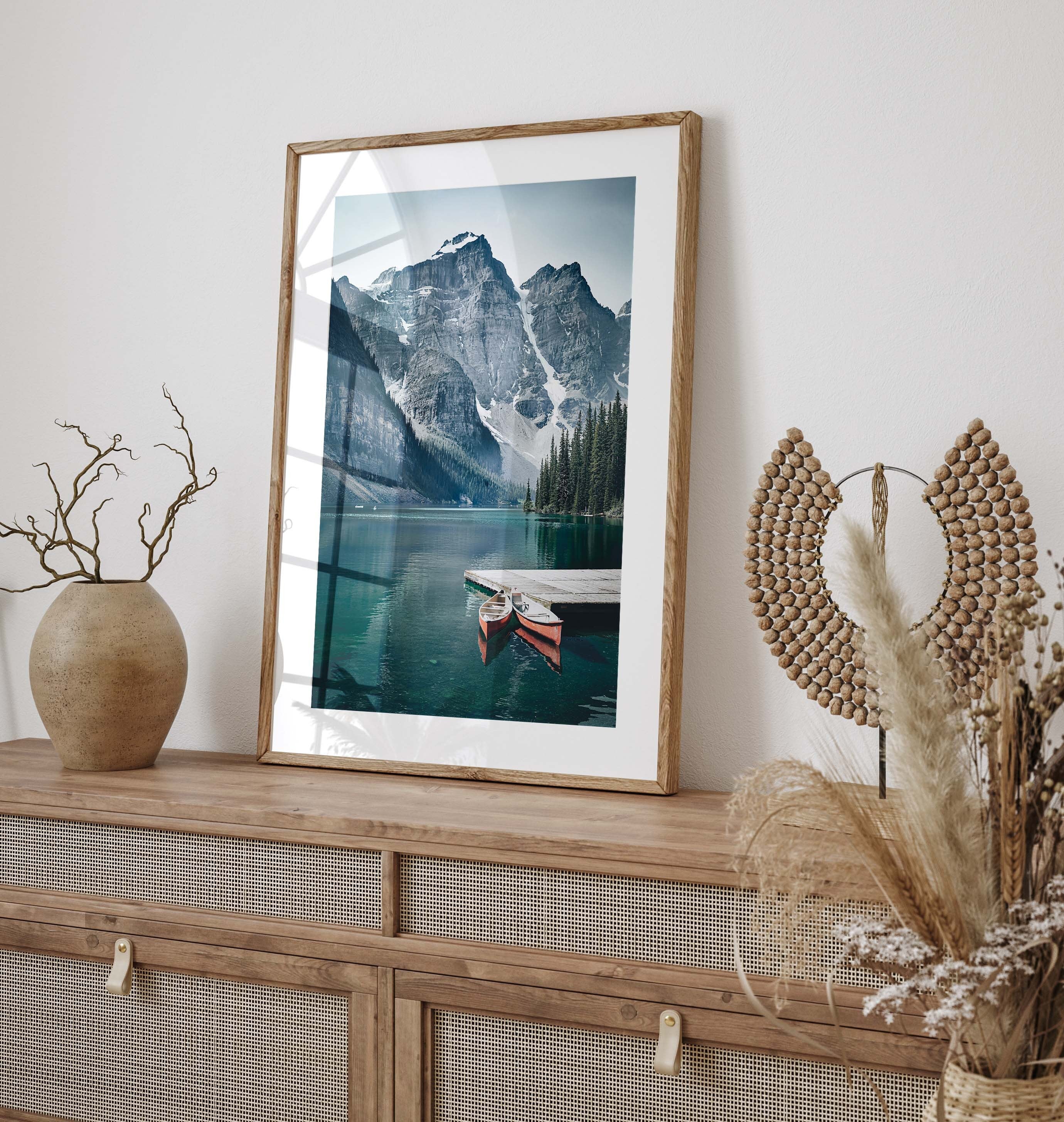 Nature poster with a photographic motif of two red canoes resting on a picturesque wooden pier on a quiet lake. On the side of the clear-blue lake, tall, ice-clad mountains are towering which is reflected beautifully on the still water surface.