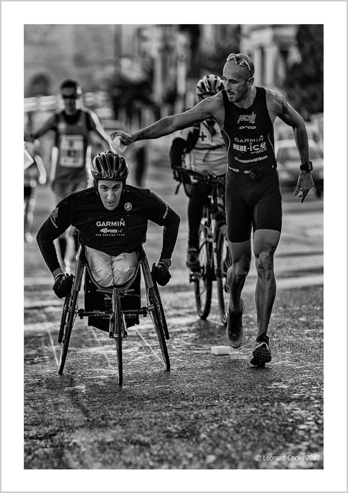 A wheelchair racer being assisted by another athlete during a race, with blurred competitors in the background. – Arteve Gallery