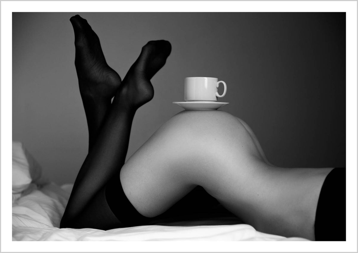 A close-up of a woman's crossed legs with a coffee cup resting on her knee, set against soft bedding. – Arteve Gallery