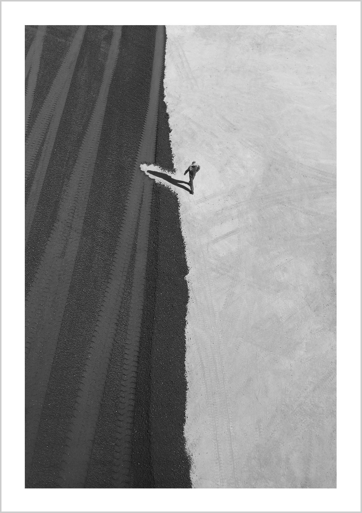An aerial view of a man walking along a dividing line in a textured landscape, showcasing strong contrasts between dark and light areas. – Arteve Gallery
