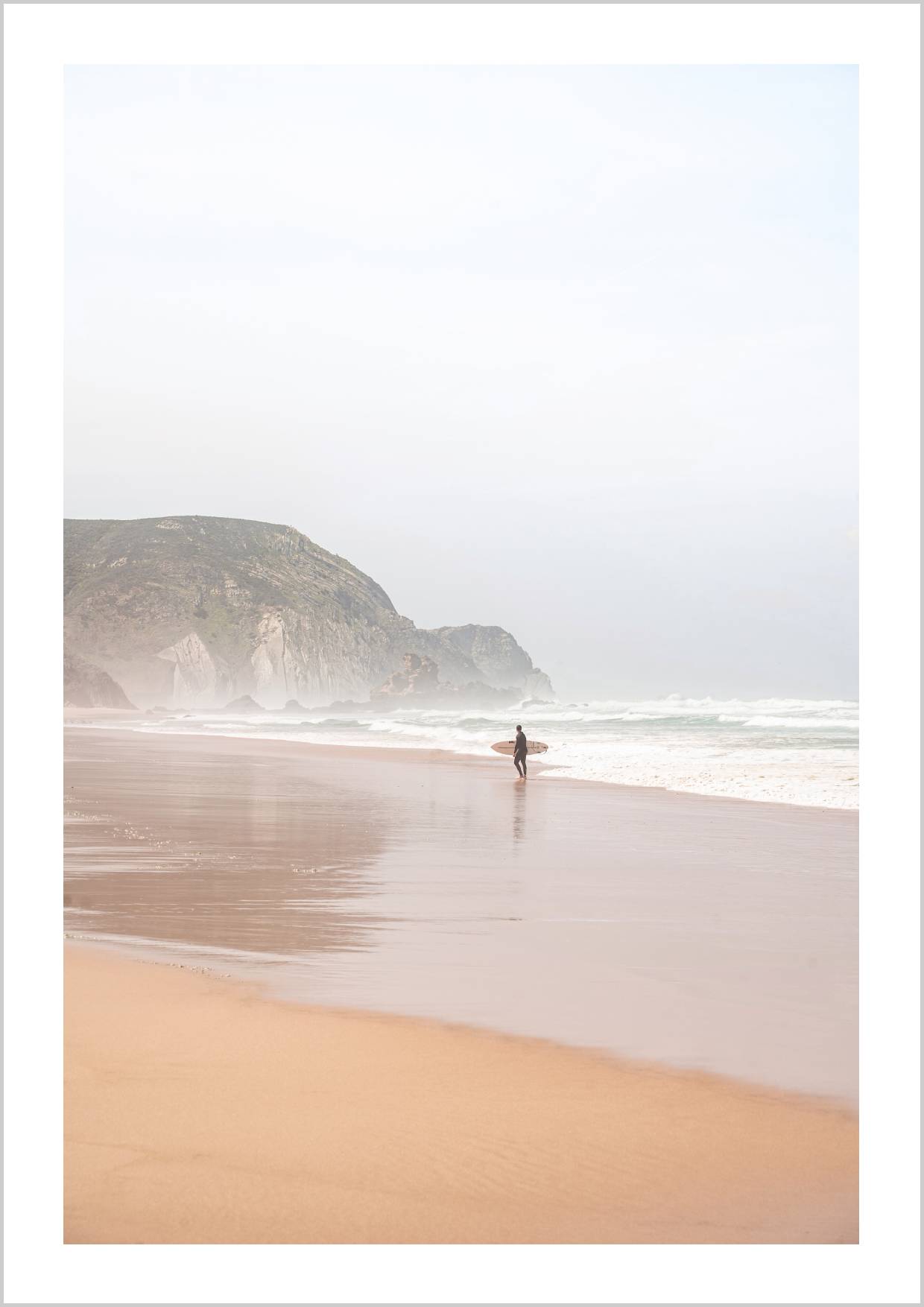 A solitary surfer walking along a misty beach with gentle waves and cliffs in the background. – Arteve Gallery